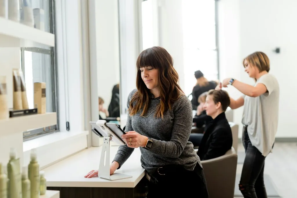 A salon owner checking appointments on a tablet at the front desk while a stylist works with a client