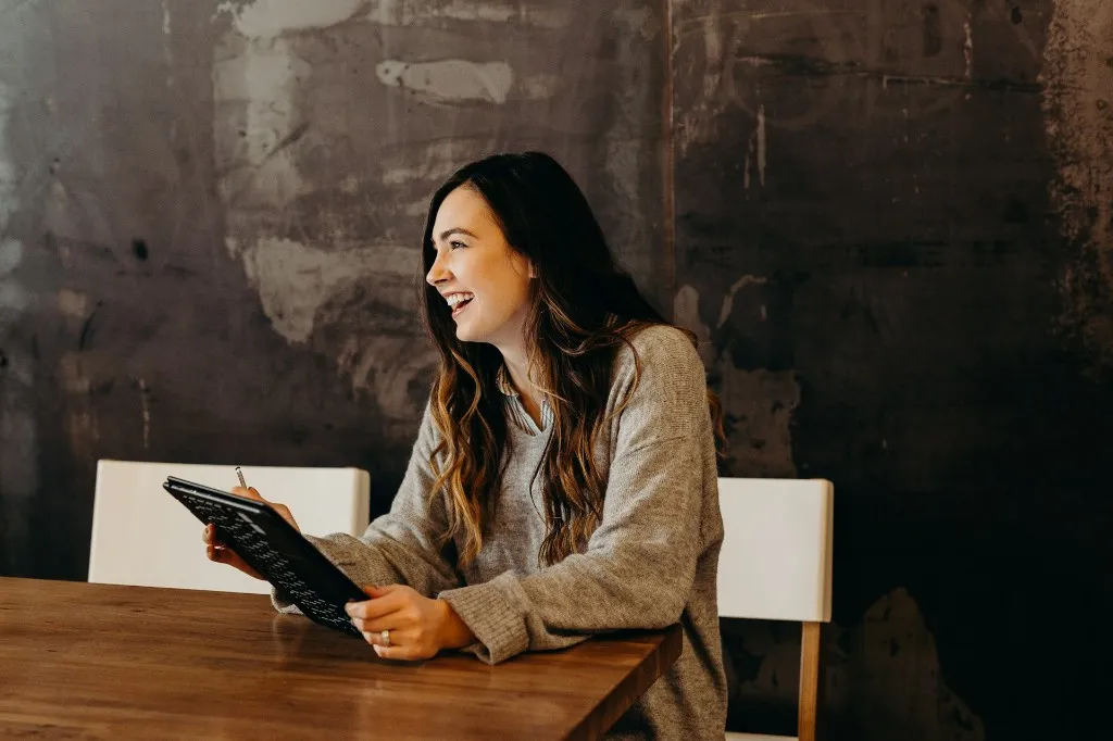 A woman smiling at a wooden table while holding a tablet and a pen